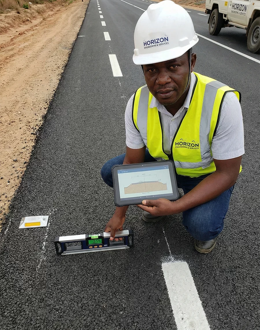A Congolese civil engineer from Horizon Engineering and Services, wearing a branded hard hat and high-vis vest, kneels on a newly paved asphalt road in Kolwezi, DR Congo. He holds a rugged tablet displaying a road cross-section diagram and uses a digital level to measure the pavement slope next to fresh lane markings.